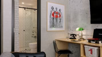 A modern bathroom with a glass shower enclosure, a toilet, and a sink. There is a framed picture on the wall, a desk with a lamp, a chair, and a vintage telephone.
