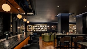 A modern bar area with a dark wood bar counter, multiple seating options including bar stools and cushioned chairs, and a display of white vases on shelves in the background. - The Anndore House, part of JdV by Hyatt (Toronto)
