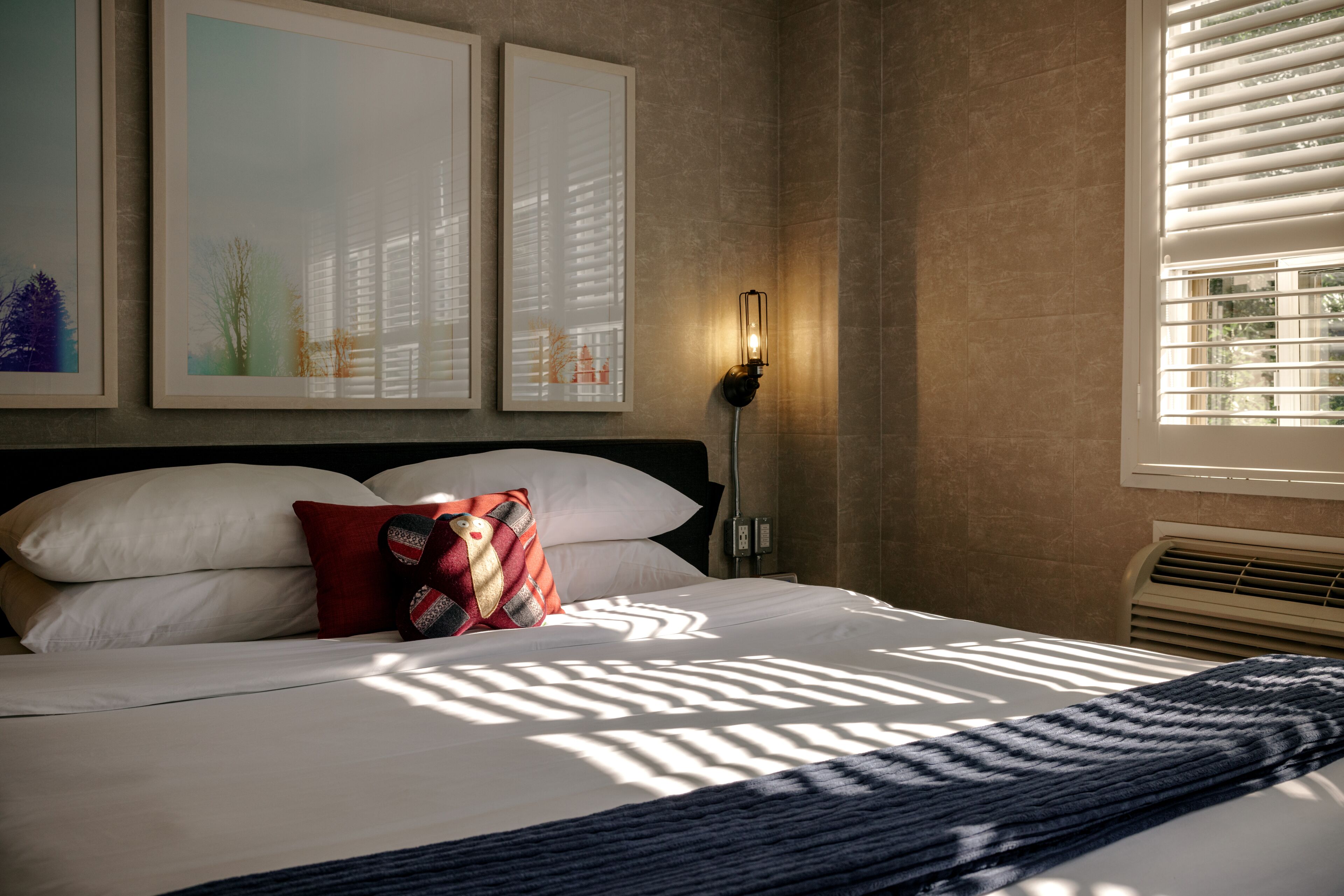 Una cama bien tendida con almohadas blancas y rojas, una manta de color azul oscuro y una lámpara de pared.
