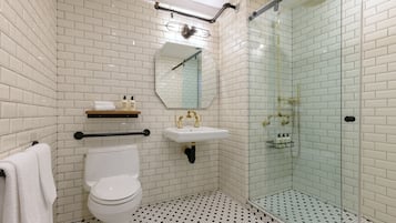 Un baño con azulejos blancos tipo metro, un inodoro, un lavabo con detalles dorados y una ducha con mampara de vidrio.
