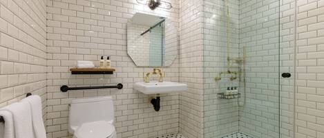 Un baño con azulejos blancos tipo metro, un inodoro, un lavabo con detalles dorados y una ducha con mampara de vidrio.