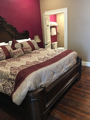 A bedroom with a wooden bed, a nightstand, a lamp, and a door leading to a bathroom. - The Kahn Hotel (Jefferson)