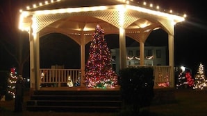 Ein Pavillon mit einem Weihnachtsbaum, der mit Lichterketten geschmückt ist, umgeben von anderen Bäumen und einem Haus im Hintergrund.