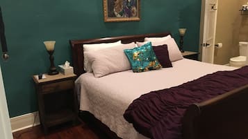 Un dormitorio con cama, mesita de noche, lámpara, un cuadro en la pared y puerta al baño.