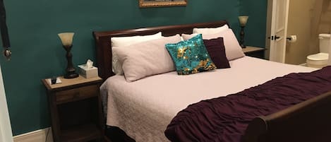 Un dormitorio con cama, mesita de noche, lámpara, un cuadro en la pared y puerta al baño.