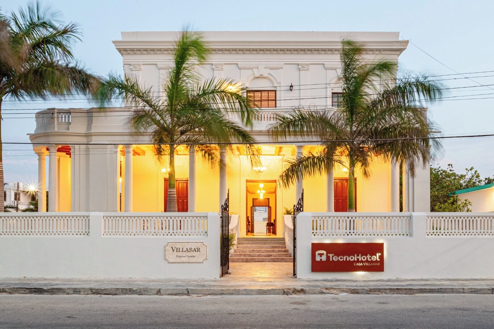 A two-story hotel with a white facade, palm trees in front, and a staircase leading to the entrance.