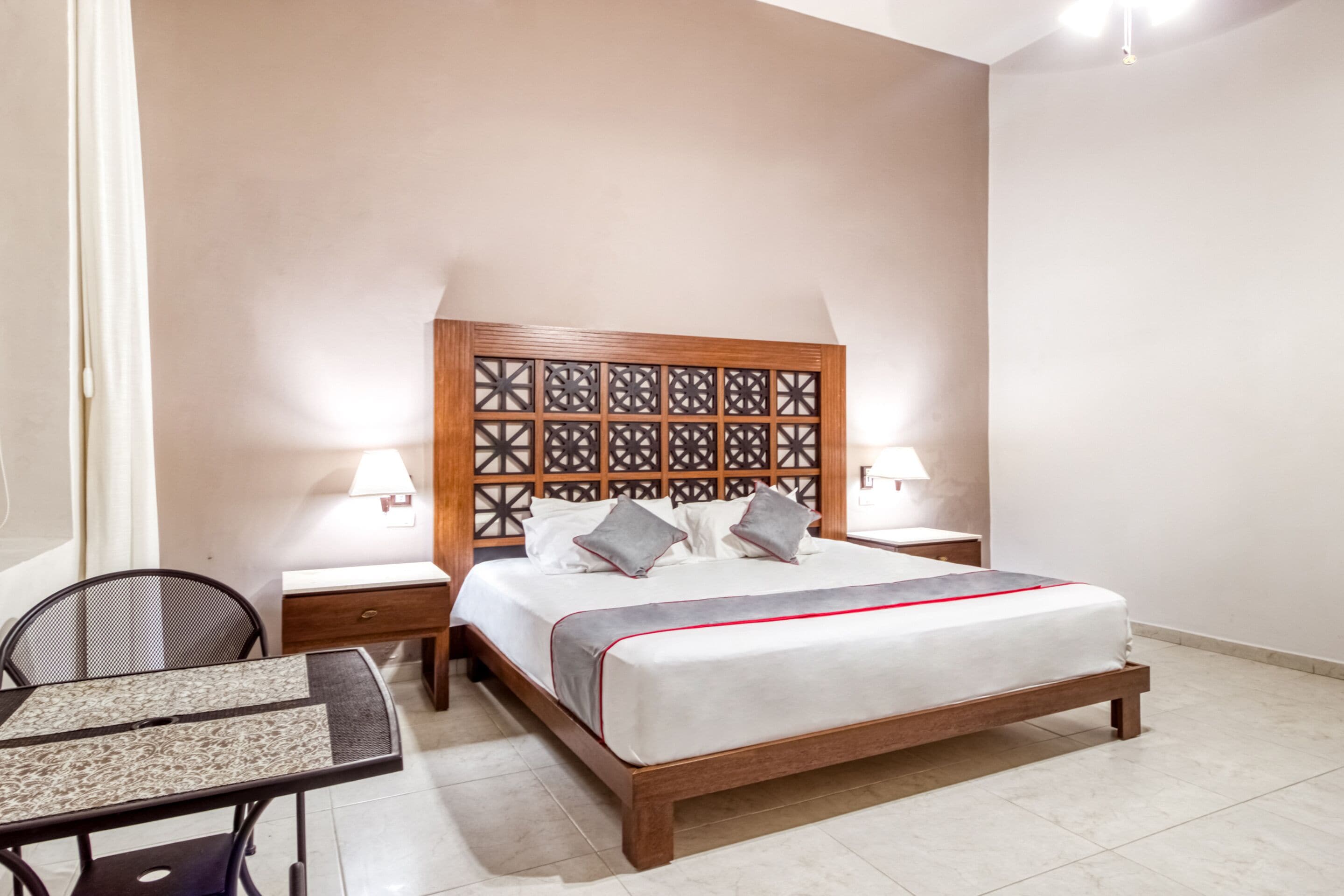 A neatly arranged bedroom with a wooden headboard, a bed with white bedding and gray pillows, a small bedside table with a lamp, and a chair at a small table.
