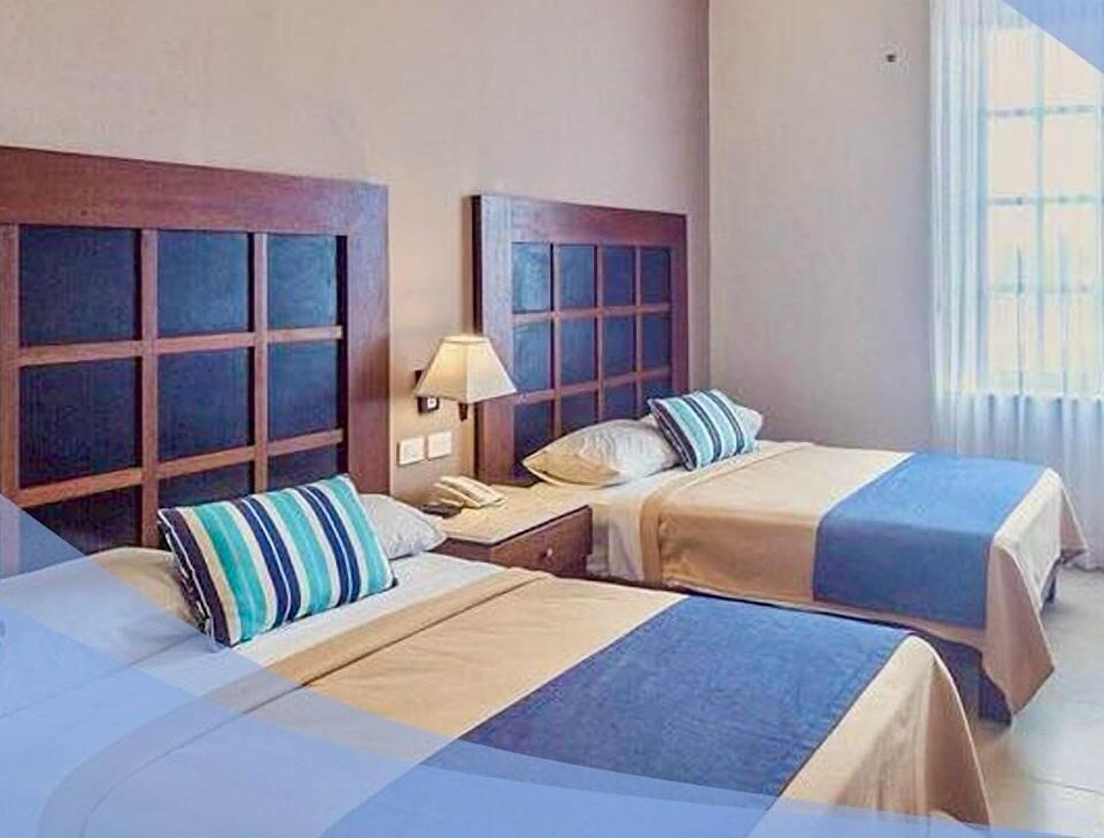 A hotel room with two beds, a nightstand, a lamp, and a window with curtains.