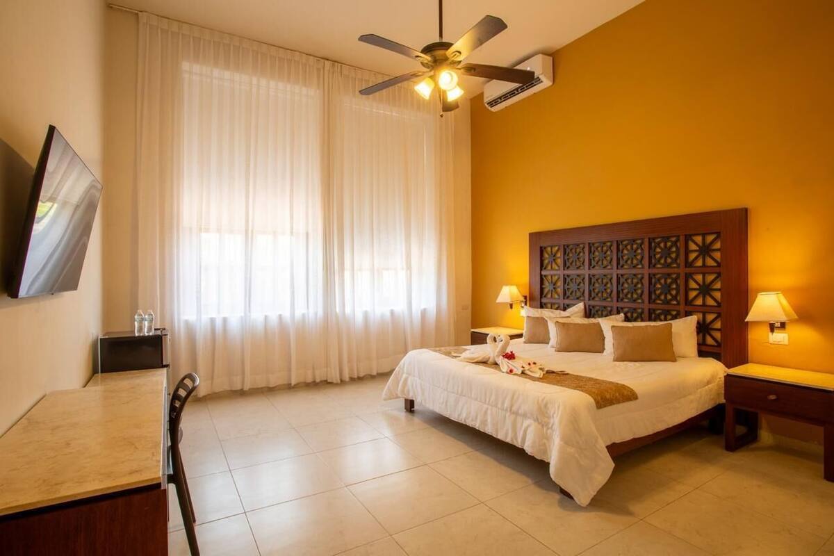 A hotel room with a bed, two bedside lamps, a ceiling fan, a TV, a desk with a chair, and a window with sheer curtains.
