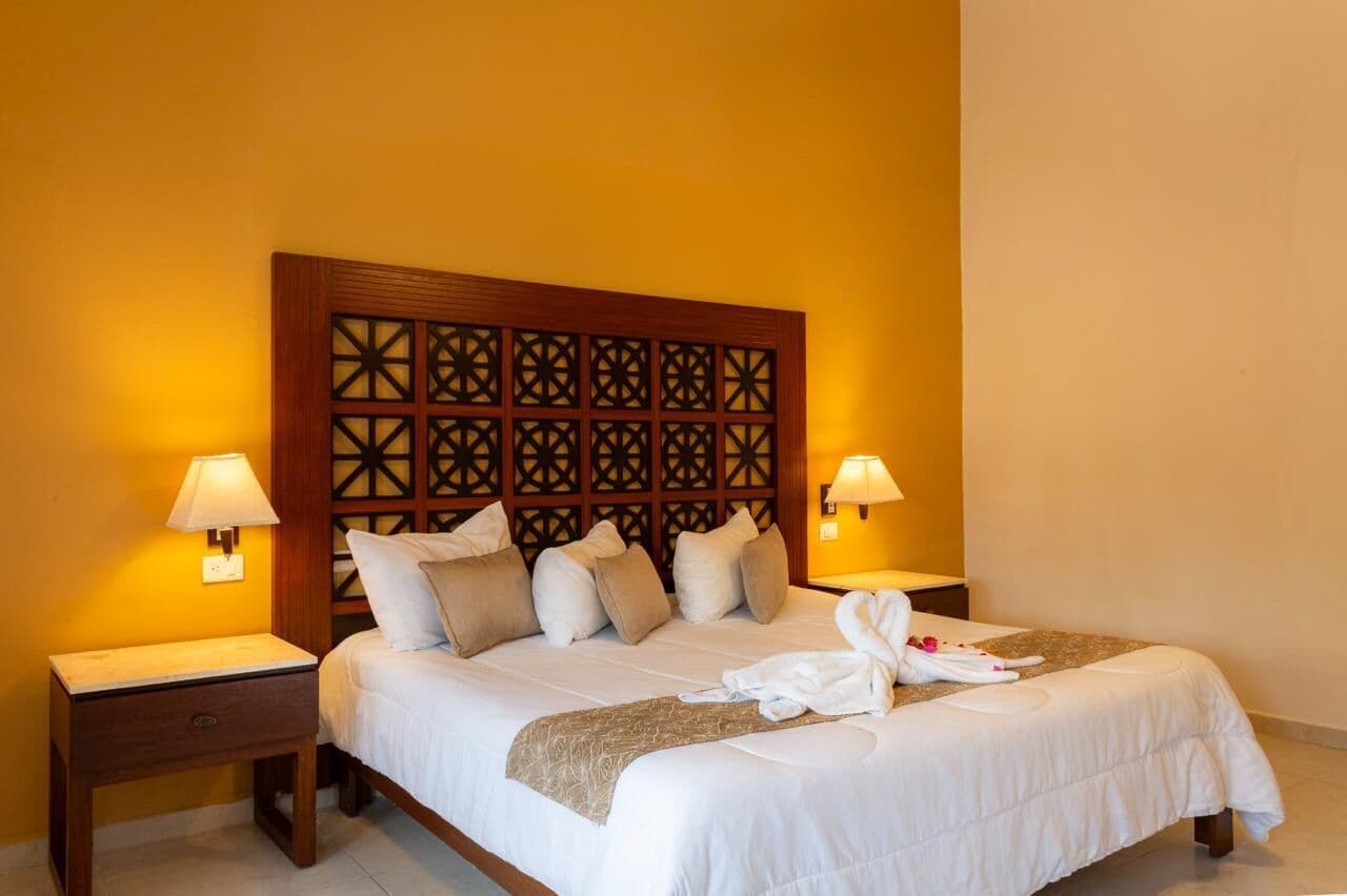 Een hotelkamer met een groot bed, twee nachtkastjes met lampen, een decoratief hoofdbord, en een net opgemaakt bed met handdoeken en een badjas.