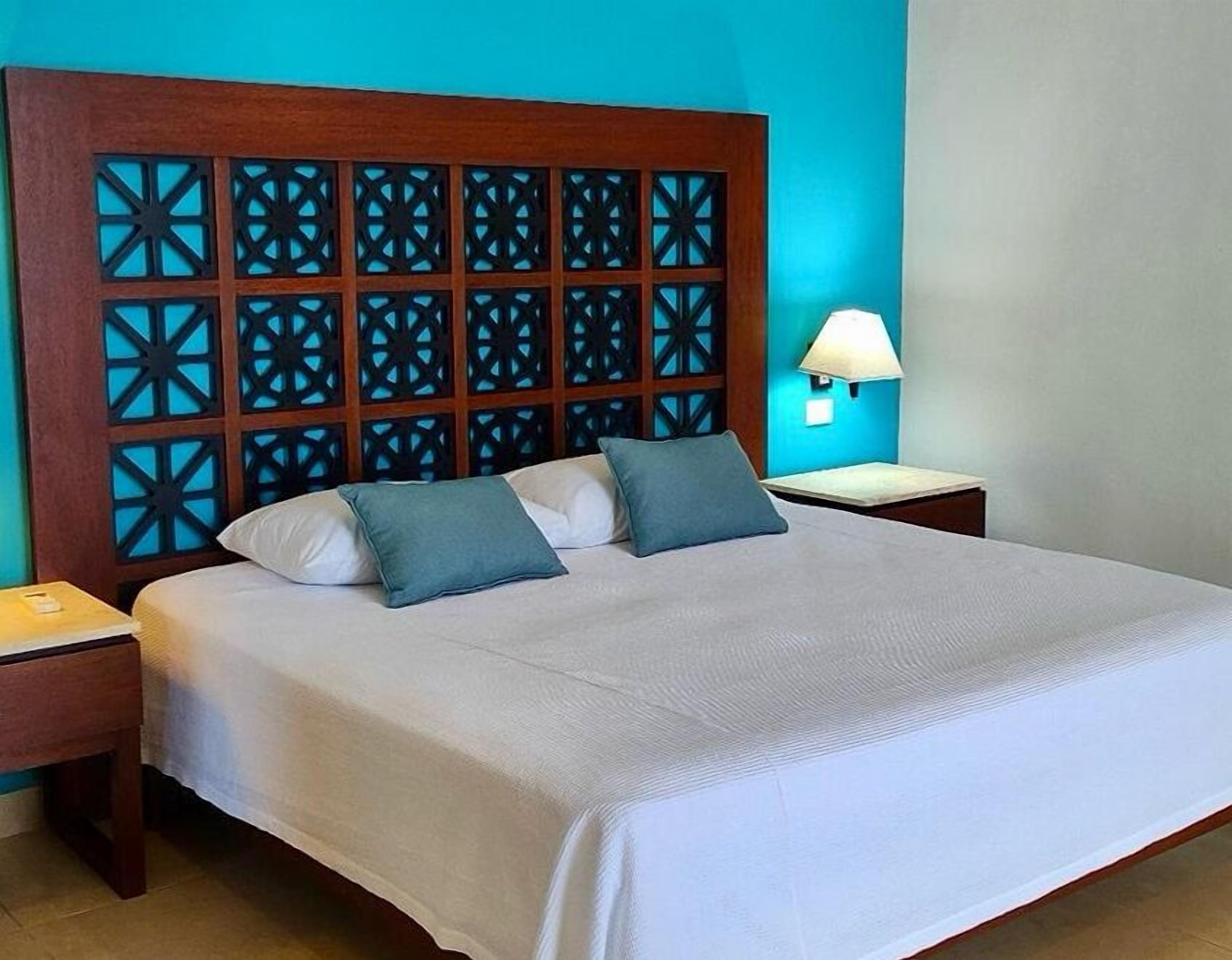 Una cama impecablemente arreglada con tablero de madera que presenta patrones geométricos, dos almohadas, una lámpara de noche y una pequeña mesita de madera.