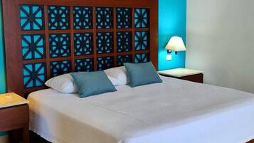 Una cama impecablemente arreglada con tablero de madera que presenta patrones geométricos, dos almohadas, una lámpara de noche y una pequeña mesita de madera.