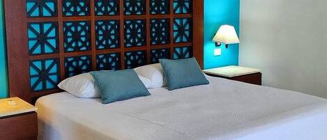 Una cama impecablemente arreglada con tablero de madera que presenta patrones geométricos, dos almohadas, una lámpara de noche y una pequeña mesita de madera.