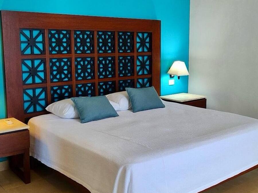 Una cama impecablemente arreglada con tablero de madera que presenta patrones geométricos, dos almohadas, una lámpara de noche y una pequeña mesita de madera.
