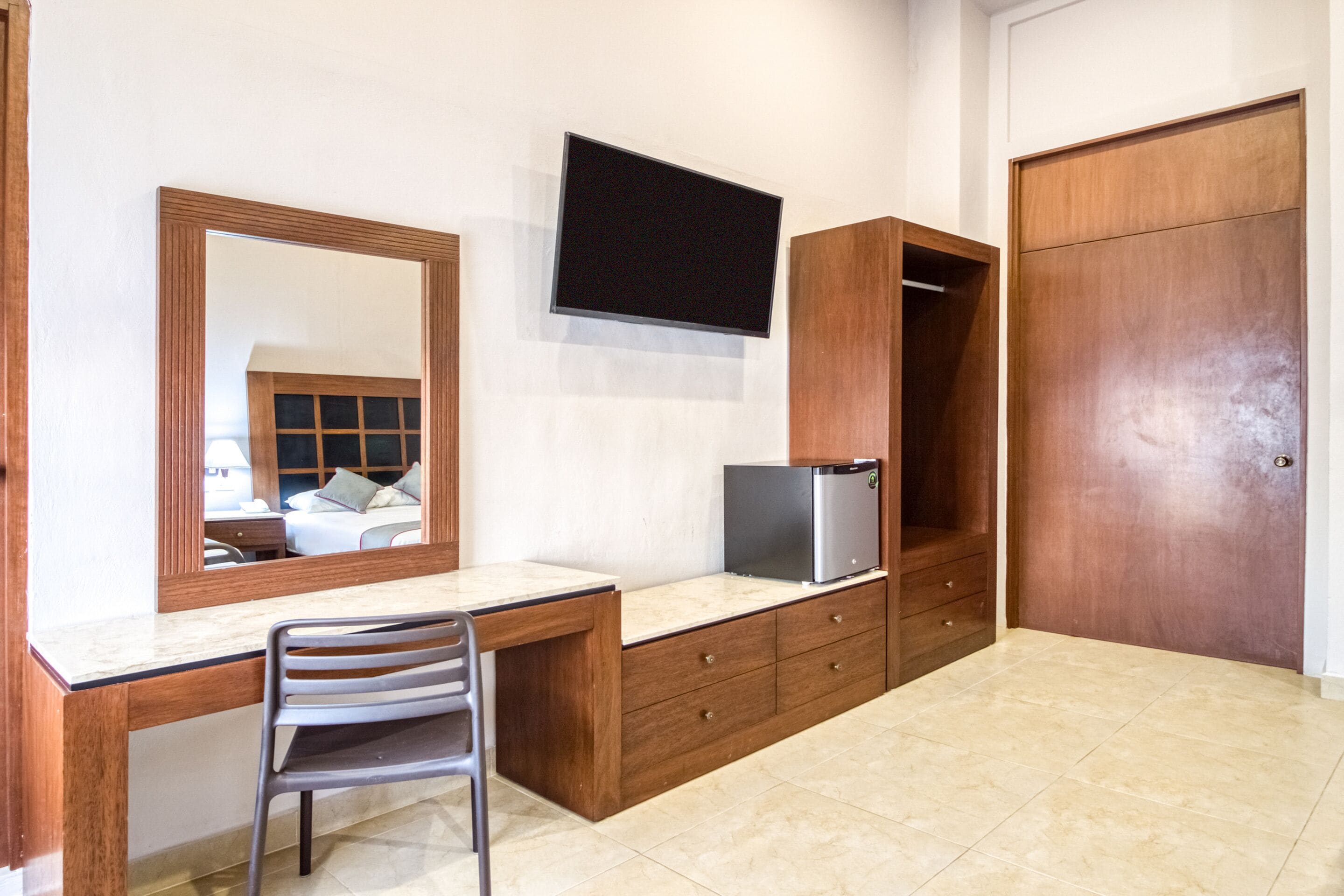 Quarto com escrivaninha de madeira, uma cadeira, televisão na parede e guarda-roupa.