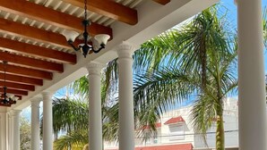 A covered outdoor area with white columns and a tiled floor, overlooking a street with palm trees and a building with a red roof. - Tecnohotel Casa Villamar (Progreso)