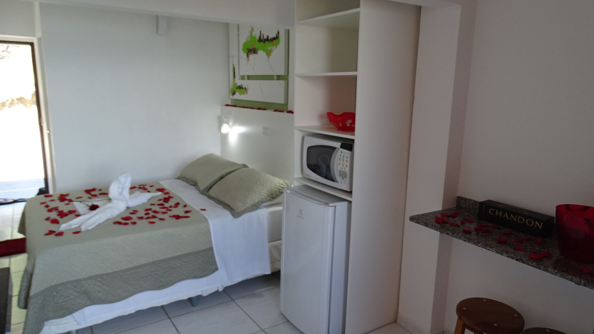 A small, compact bedroom with a bed, a white refrigerator, a microwave, and a small shelf with a red object.