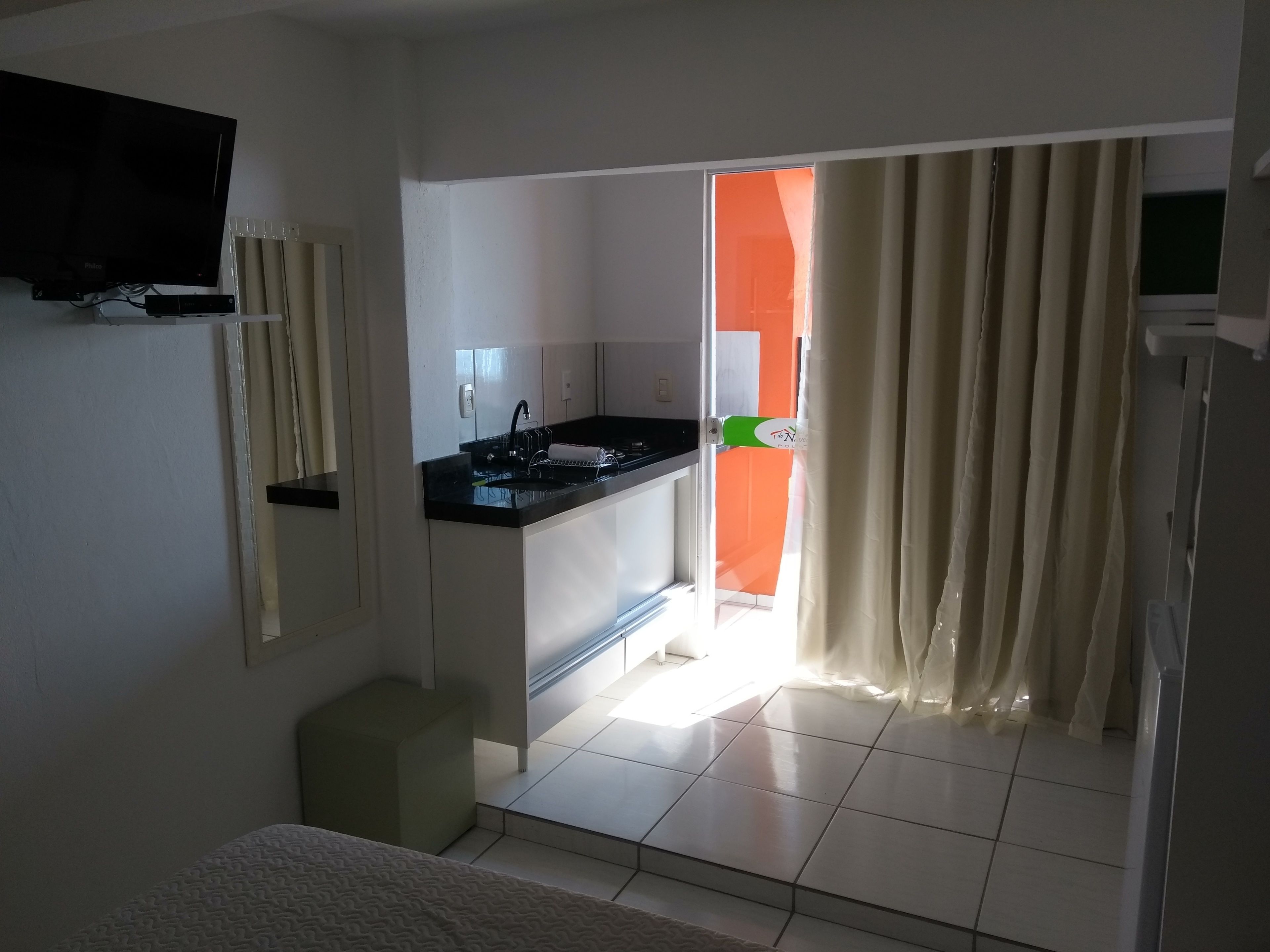 Cocina compacta con encimera negra, lavamanos y un taburete pequeño. Un televisor está montado en la pared, y una puerta corrediza da a una habitación luminosa con paredes anaranjadas.