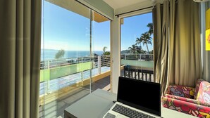 Una habitación con una computadora portátil sobre una mesa, un balcón con vistas al océano y una silla colorida.