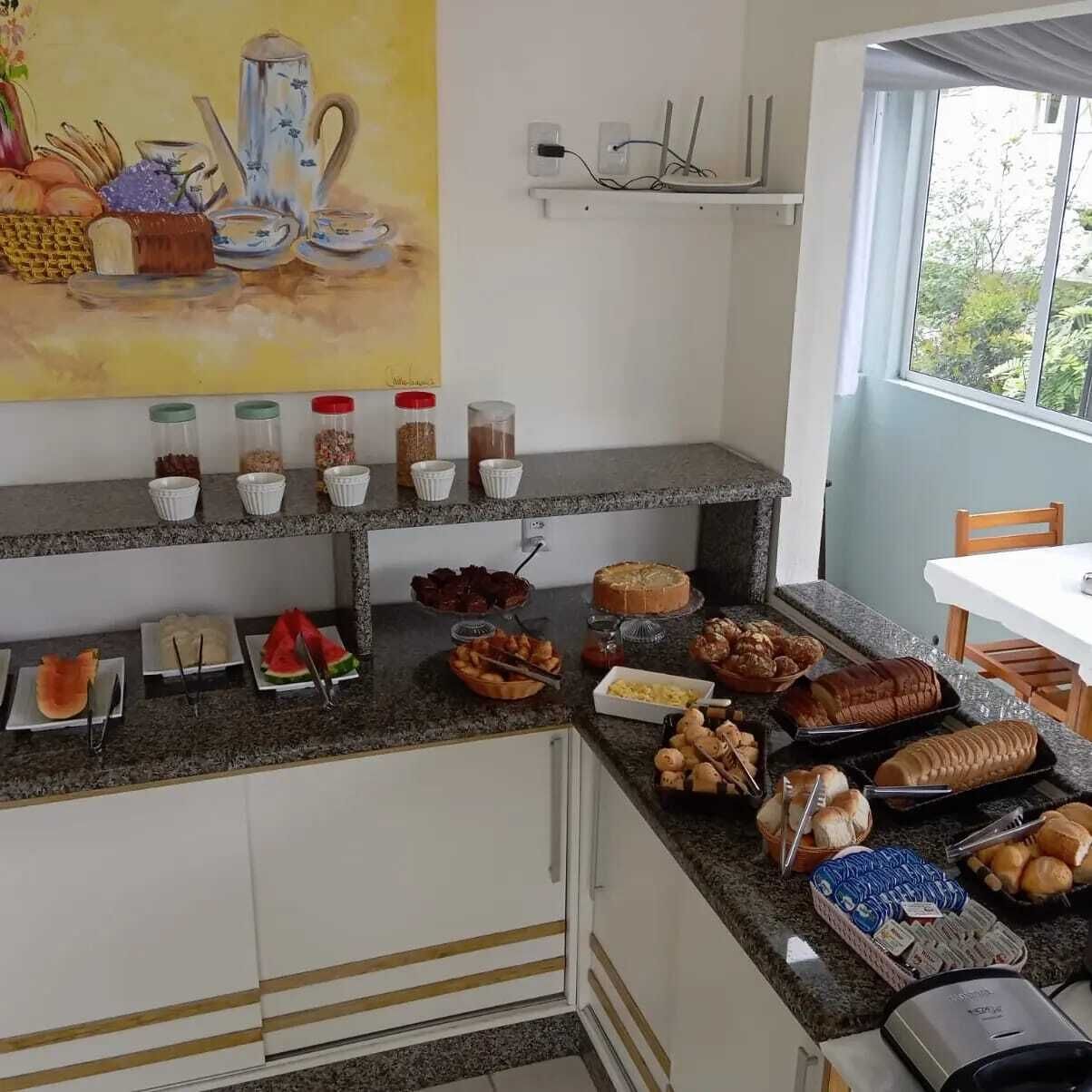 A kitchen with a variety of food items on the counter, including bread, pastries, and condiments. There is a painting on the wall above the counter, and a window providing a view of the outdoors.