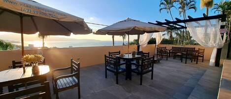 An outdoor patio with tables and chairs, umbrellas, and a view of the sea.
