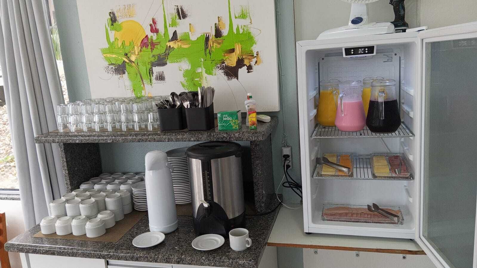 A kitchen area with a refrigerator stocked with beverages, a coffee station with cups and a coffee maker, and a wall with a colorful abstract painting.
