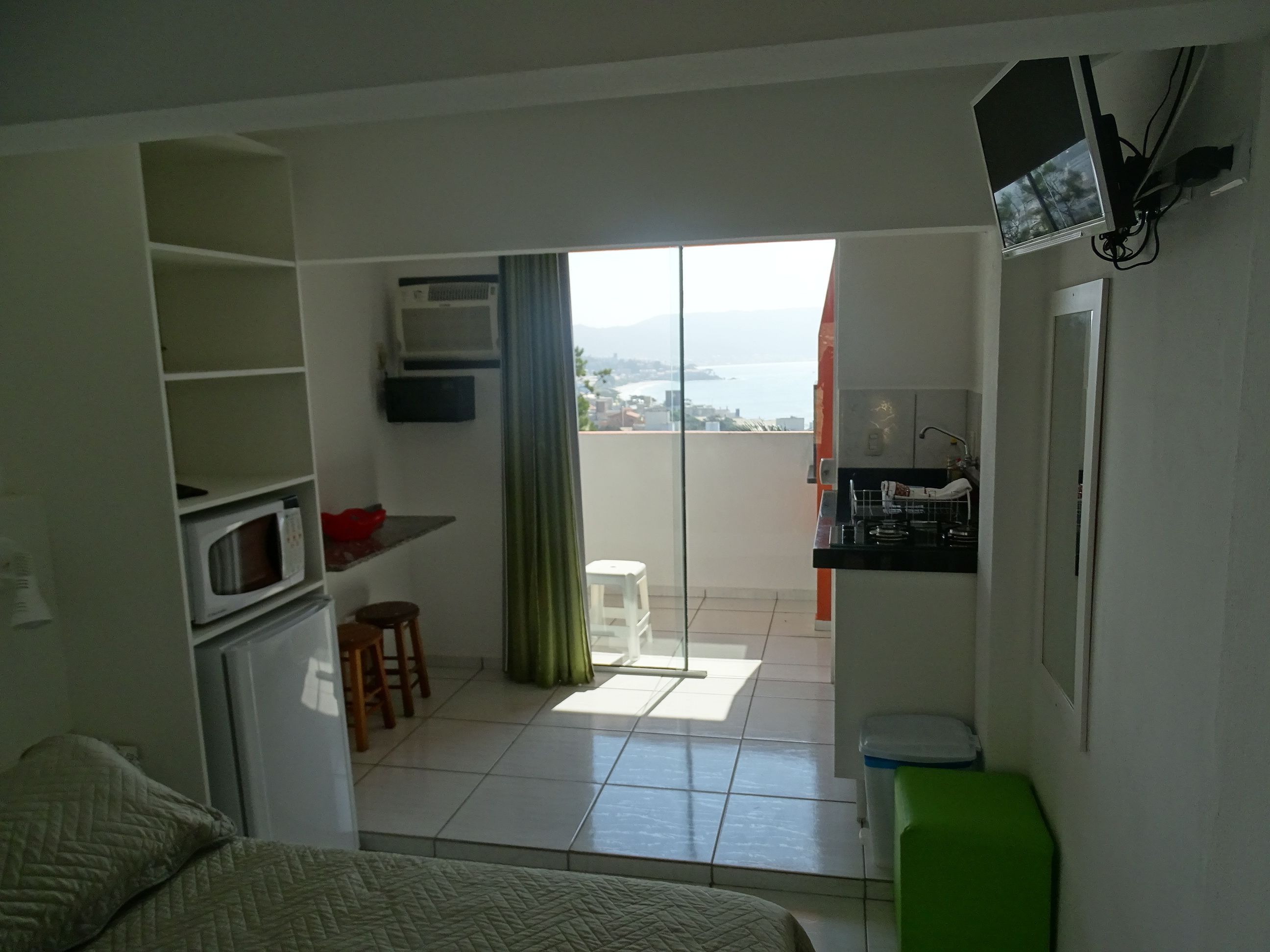 A compact hotel room with a balcony, a kitchenette, a bed, and a small seating area.