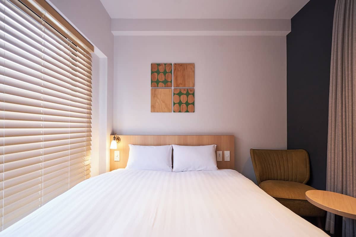 Quarto de hotel com uma cama grande, cabeceira de madeira, luminária fixada na parede, mesa redonda pequena e uma cadeira.