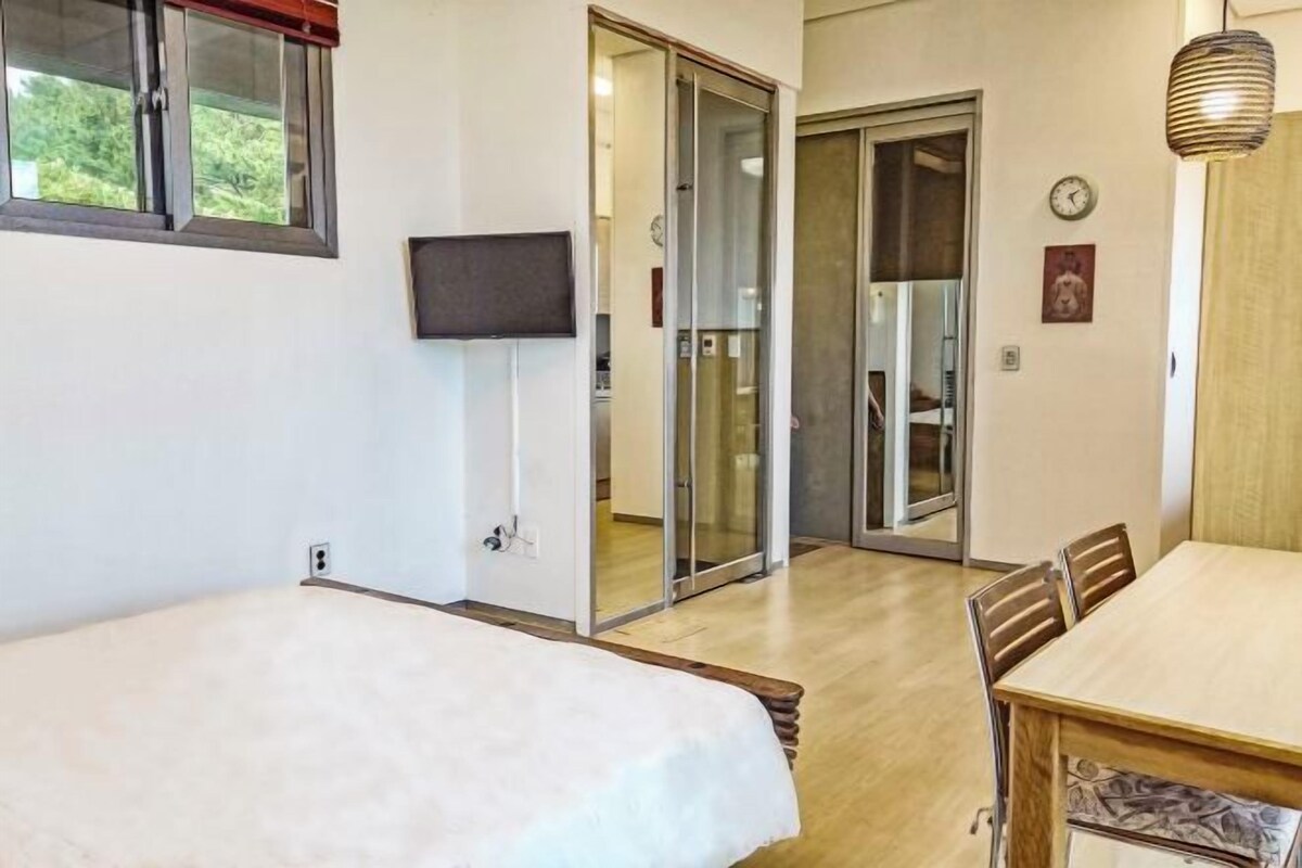 Quarto com cama, televisão, mesa de jantar e porta de vidro deslizante para um banheiro.