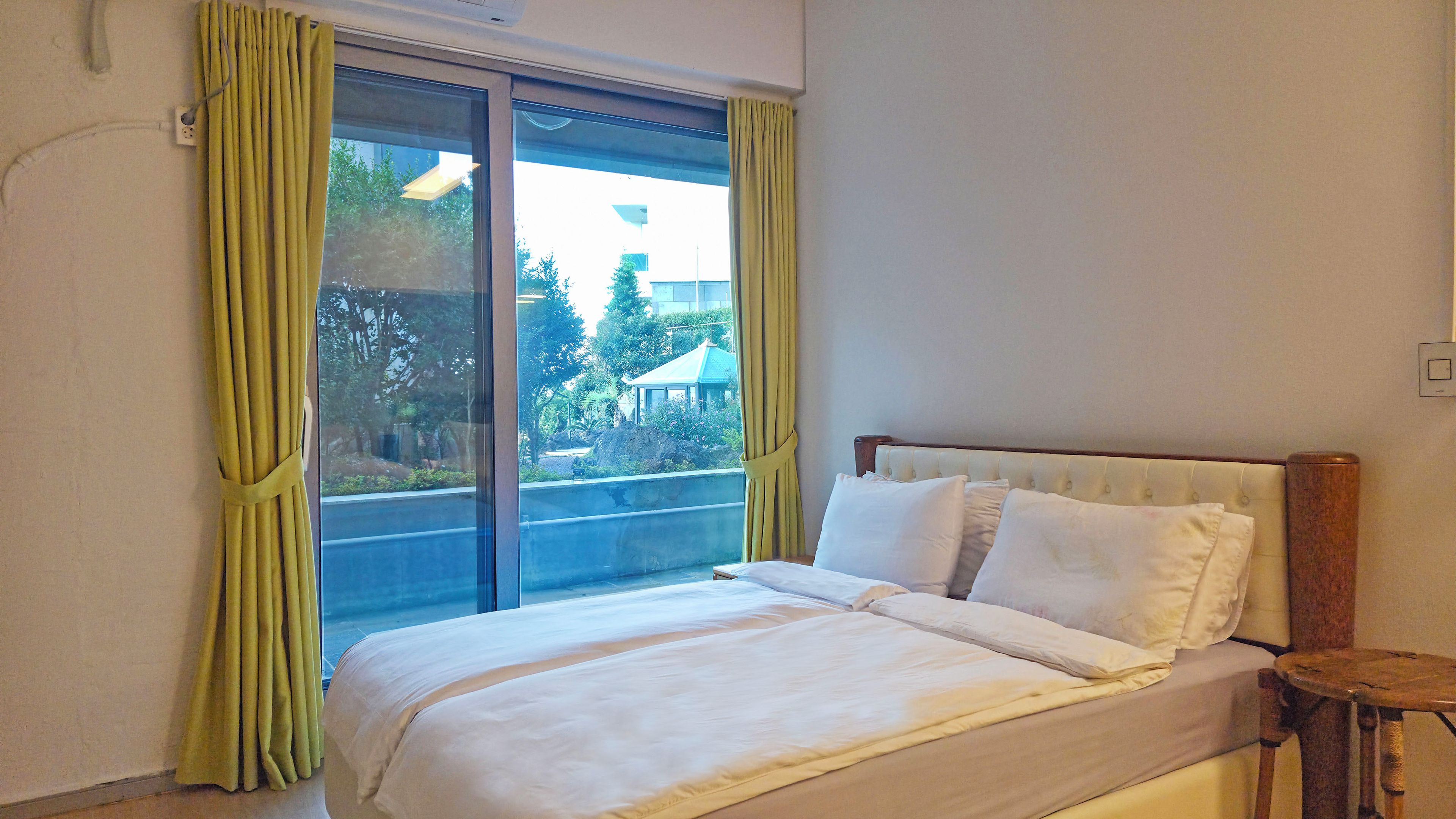 A hotel room with a bed, a small wooden side table, and a view of greenery through a window with yellow curtains.