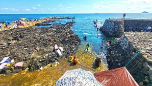 Zona costera rocosa con agua azul cristalina, gente nadando y tomando el sol, y un muro de piedra en primer plano.