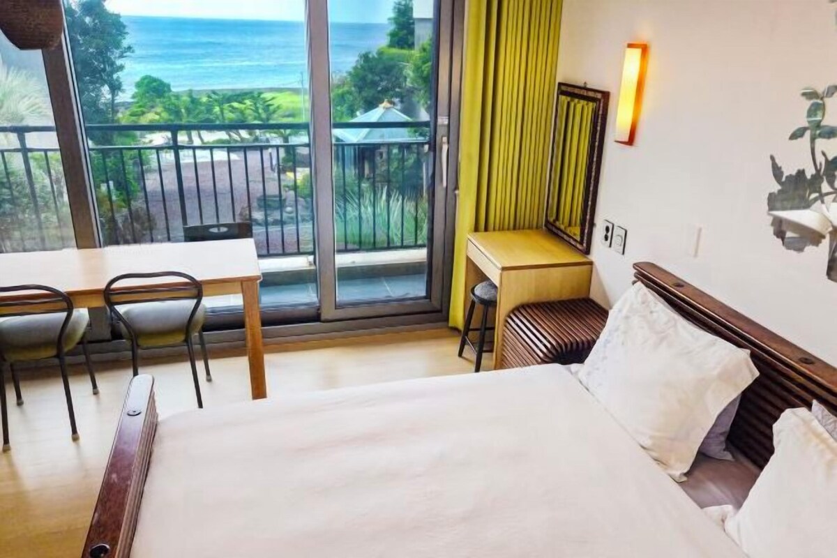 A hotel room with a bed, a desk, a chair, a window with a view of the ocean, and a wall-mounted light.