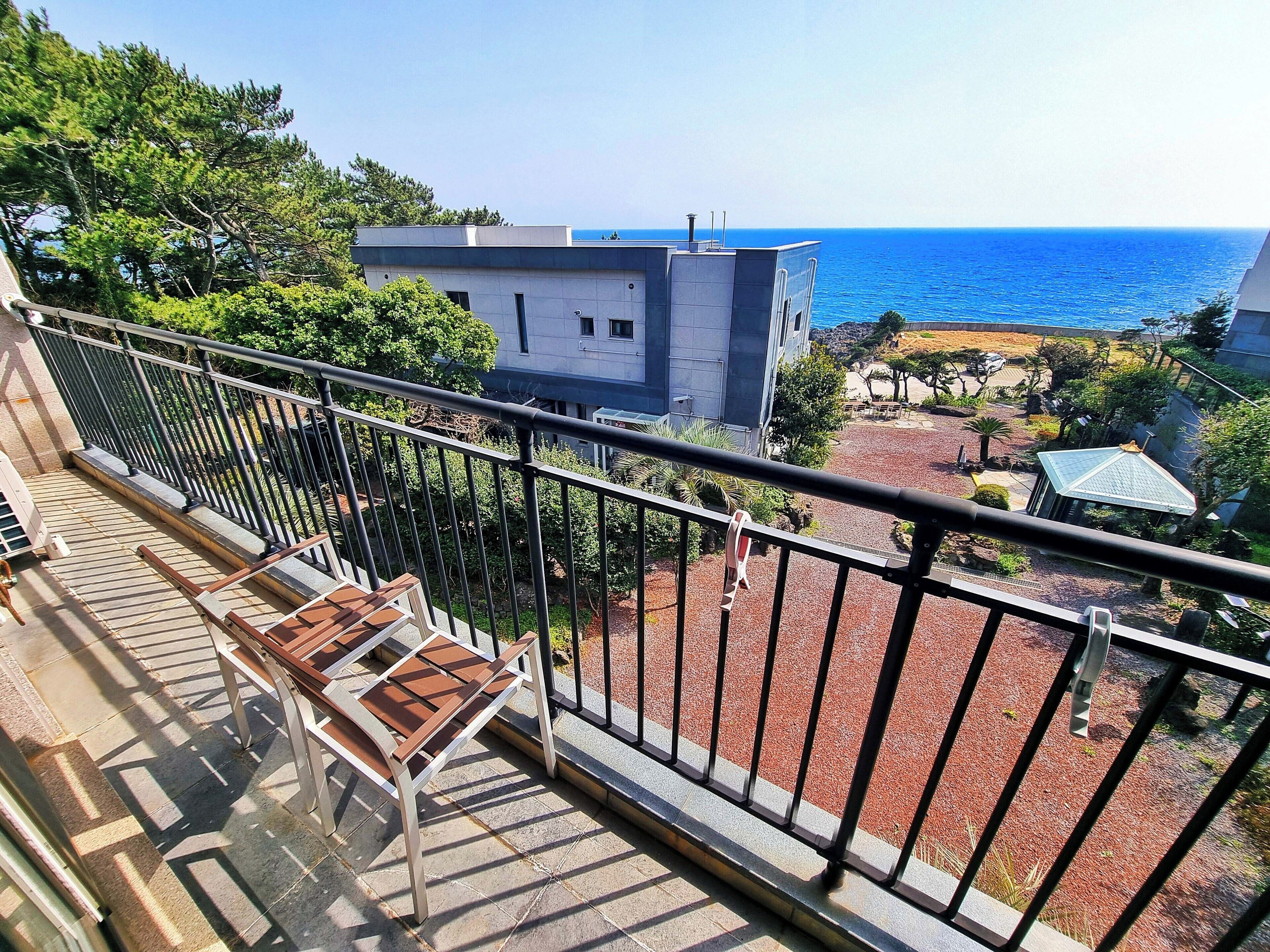 Un balcón con vista al mar, un edificio al fondo y un jardín con una banca.