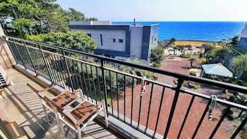 Un balcón con vista al mar, un edificio al fondo y un jardín con una banca.