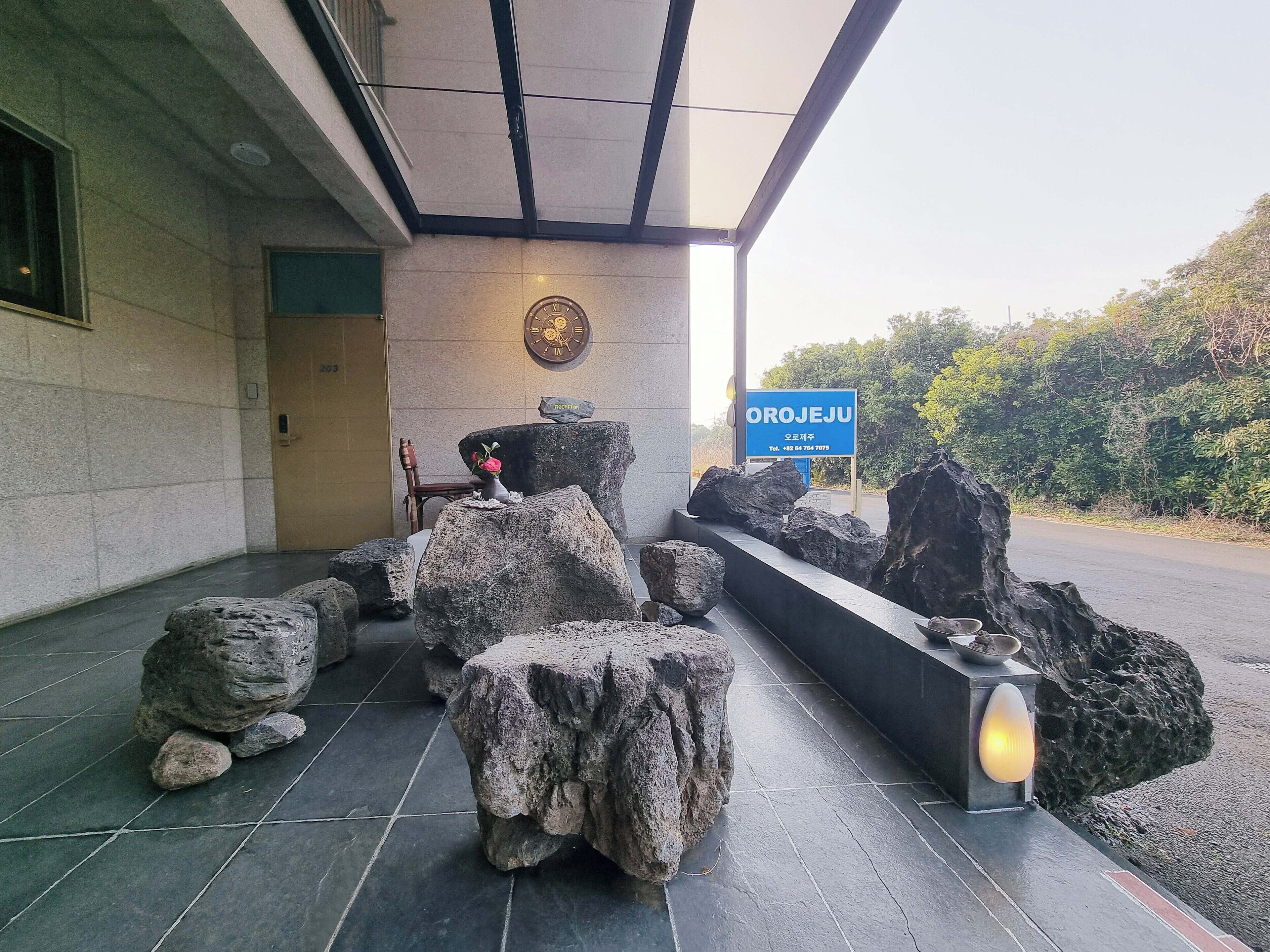 Zona con bancos de piedra, un reloj, un cartel y un camino que lleva a la entrada de un edificio.