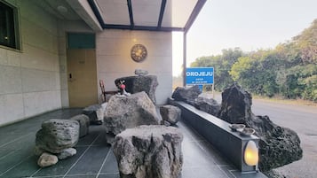 Zona con bancos de piedra, un reloj, un cartel y un camino que lleva a la entrada de un edificio.