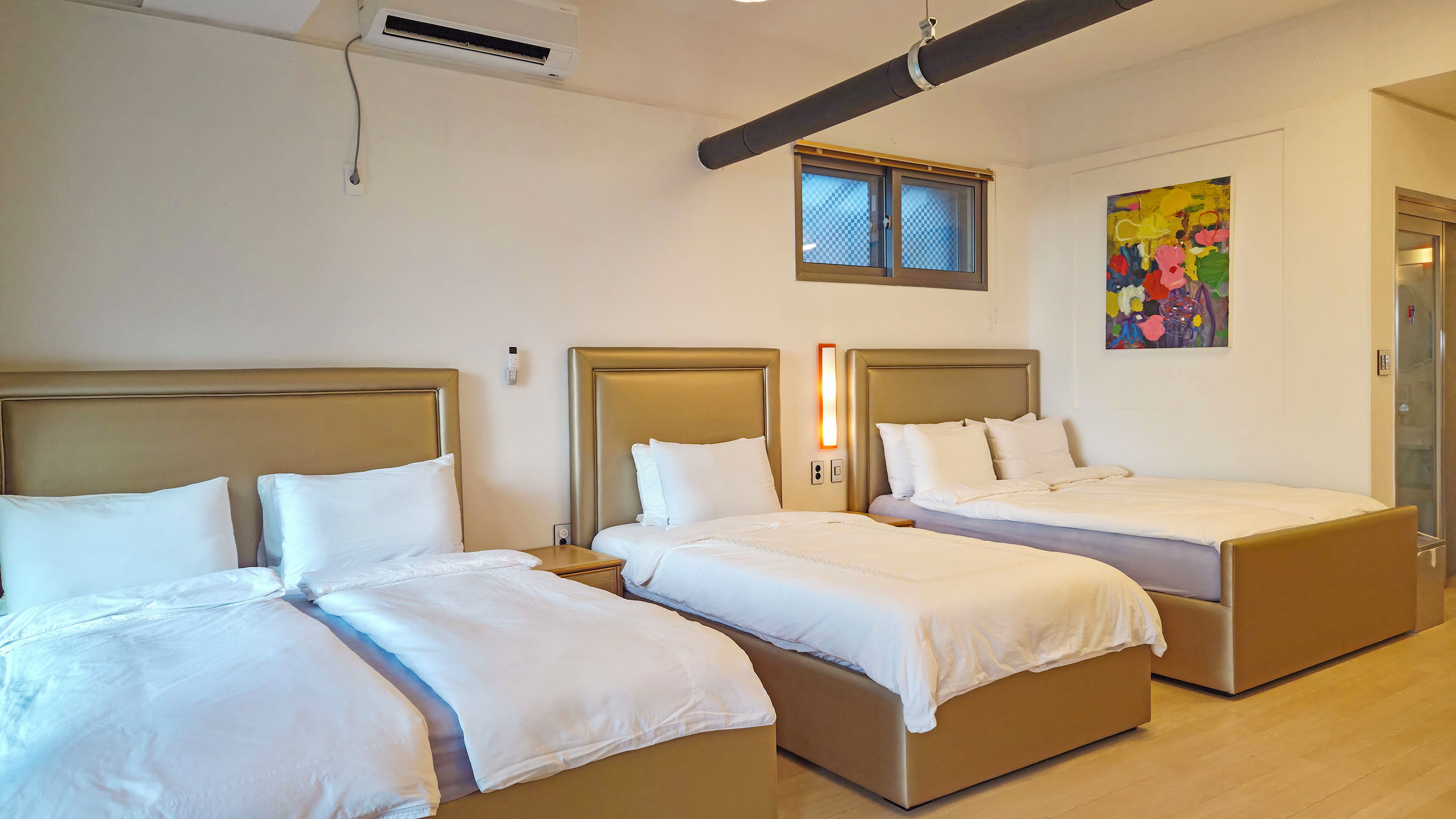 A hotel room with three beds, a window, a painting, and an air conditioning unit.
