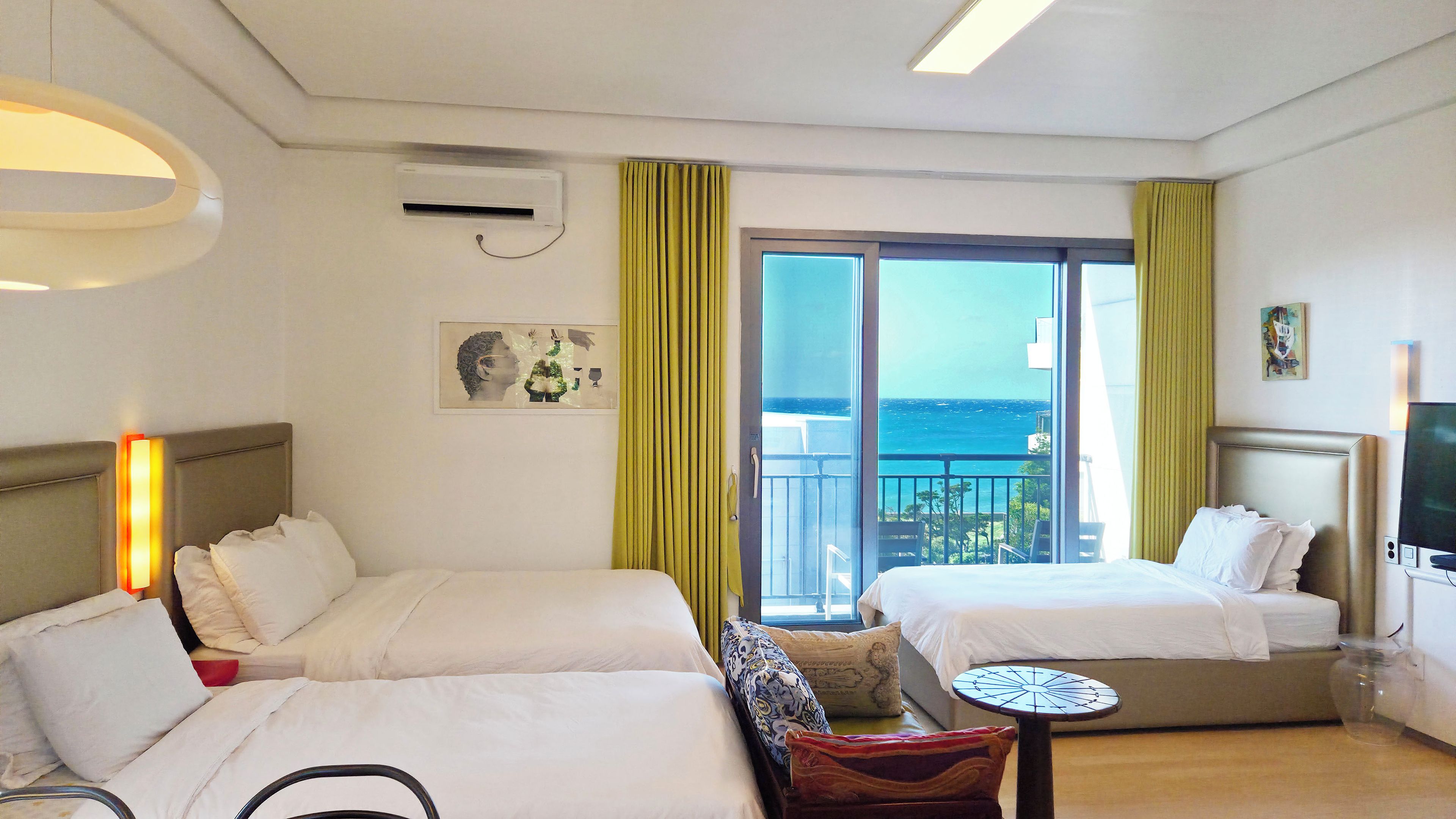 A hotel room with two beds, a small table, a chair, a TV, and a balcony with a view of the sea.