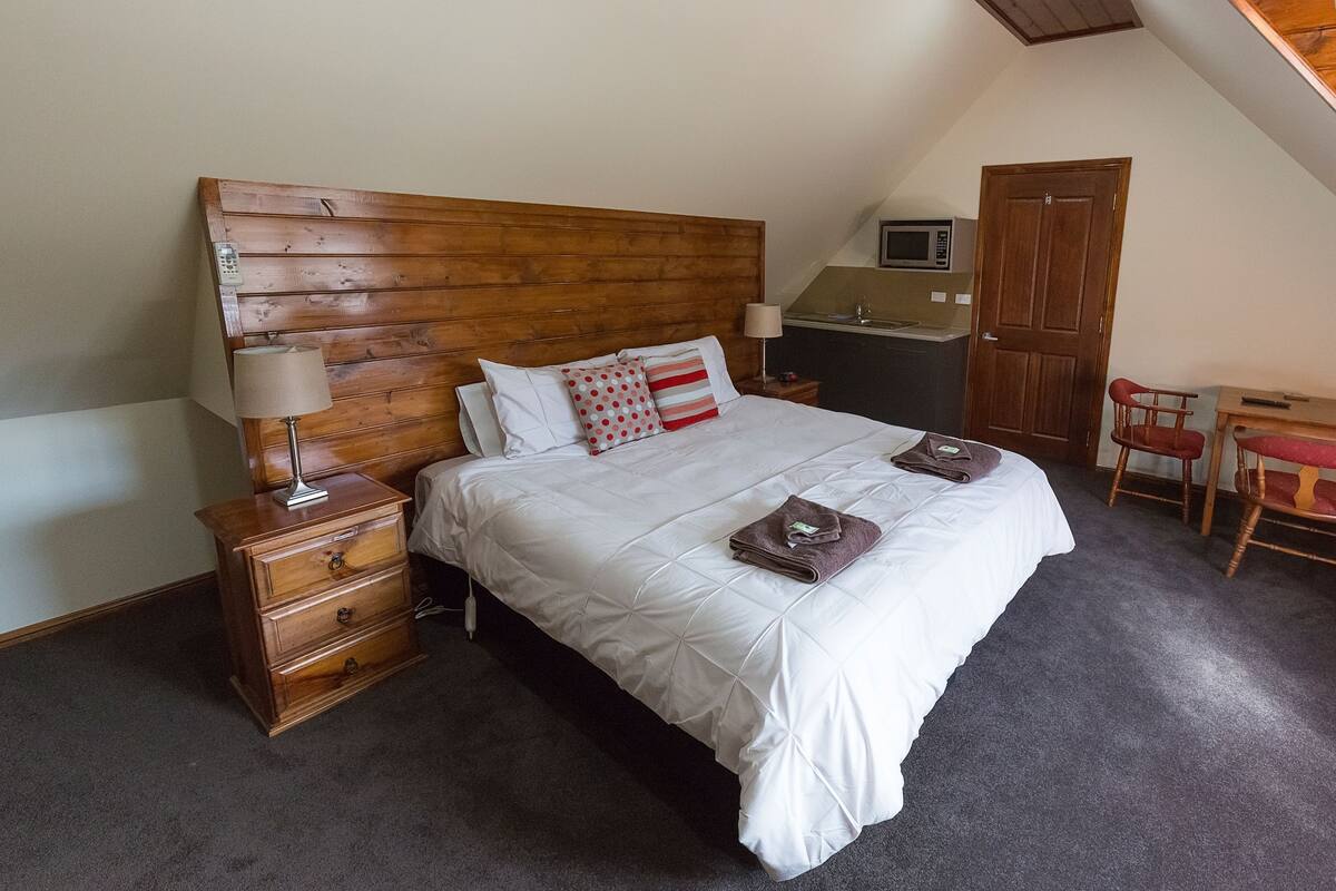 Une chambre avec une tête de lit en bois, un lit avec une literie blanche, une table de chevet avec une lampe, un bureau avec une chaise et un micro-ondes posé sur la commode.