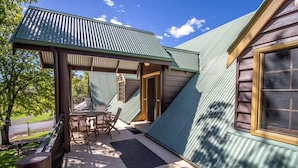A covered outdoor area with a table and chairs, a wooden deck, and a building with a green roof. - Gold Mine Guesthouse (Nundle)
