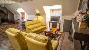 A cozy living room with a yellow sofa set, a coffee table, and a TV. There are two windows with blinds, a lamp, and a fireplace. - Gold Mine Guesthouse (Nundle)