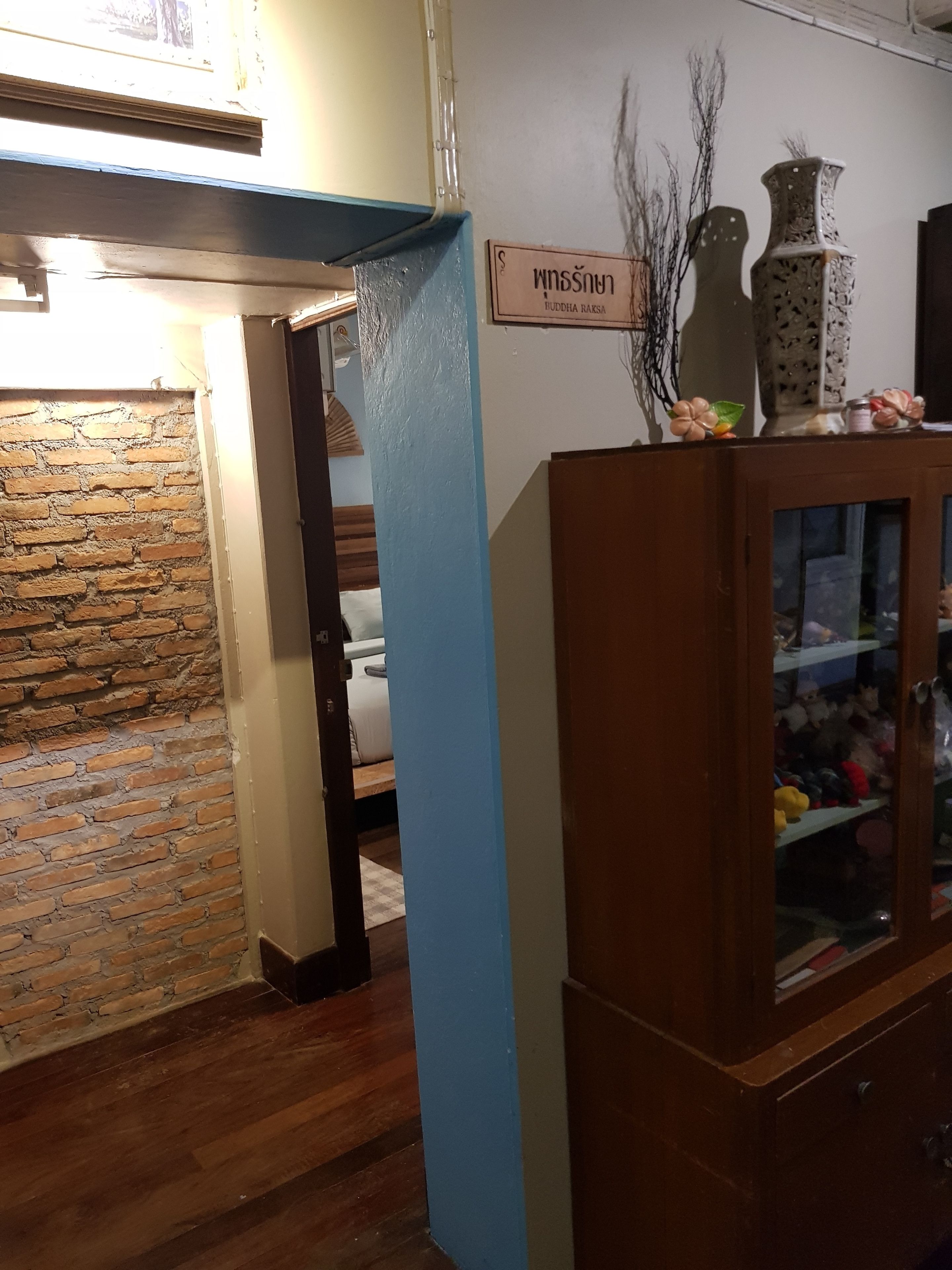 A room with a brick wall, a blue painted pillar, a wooden cabinet with glass doors, and a sign with Thai text.