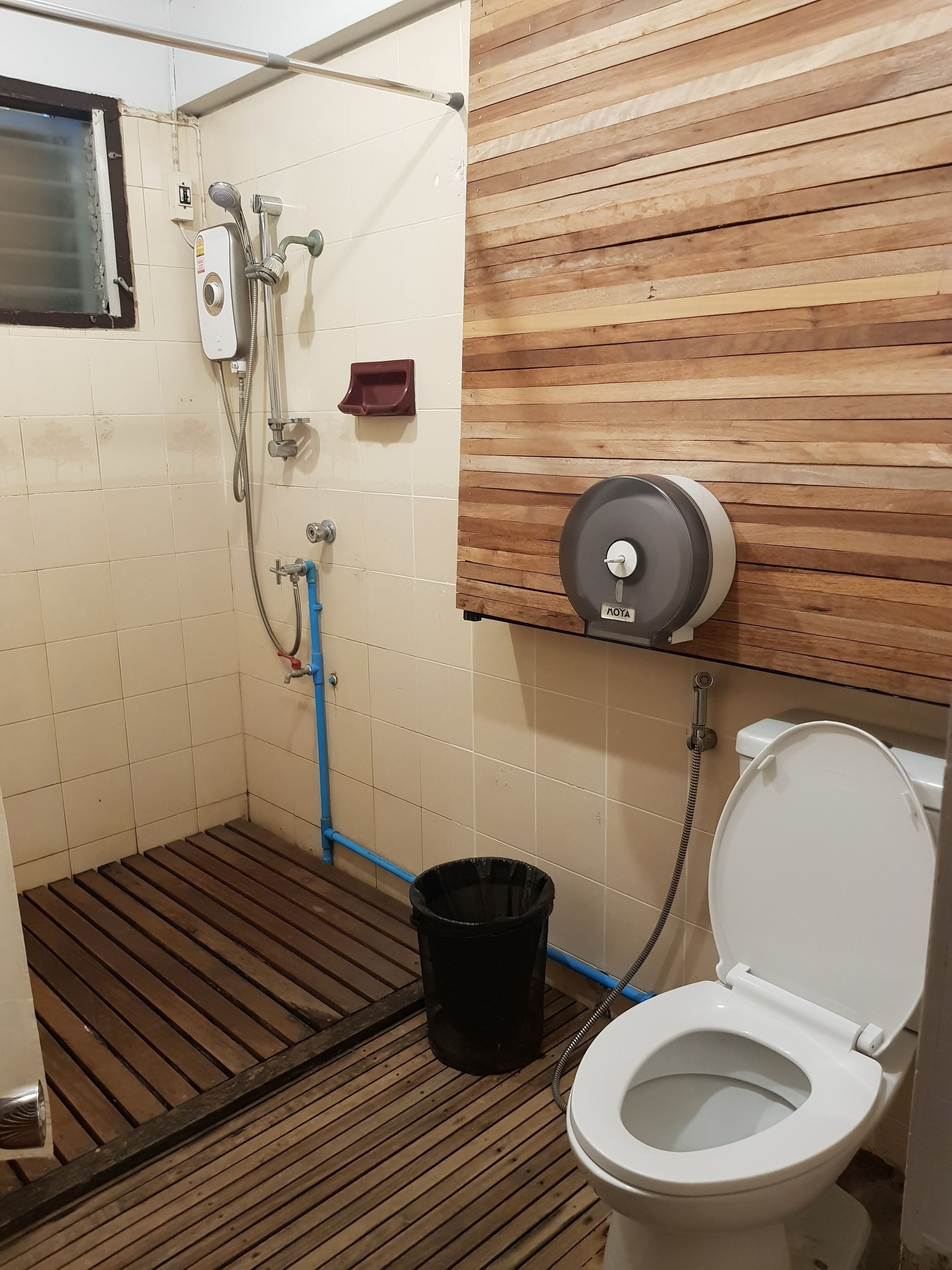 A small bathroom with a wooden floor, a white toilet, a shower with a hose, and a trash bin.