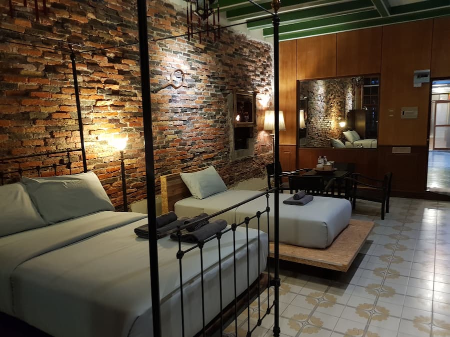 Quarto de hotel com parede de tijolos, cama com estrutura de metal, sofá, mesa pequena e espelho.