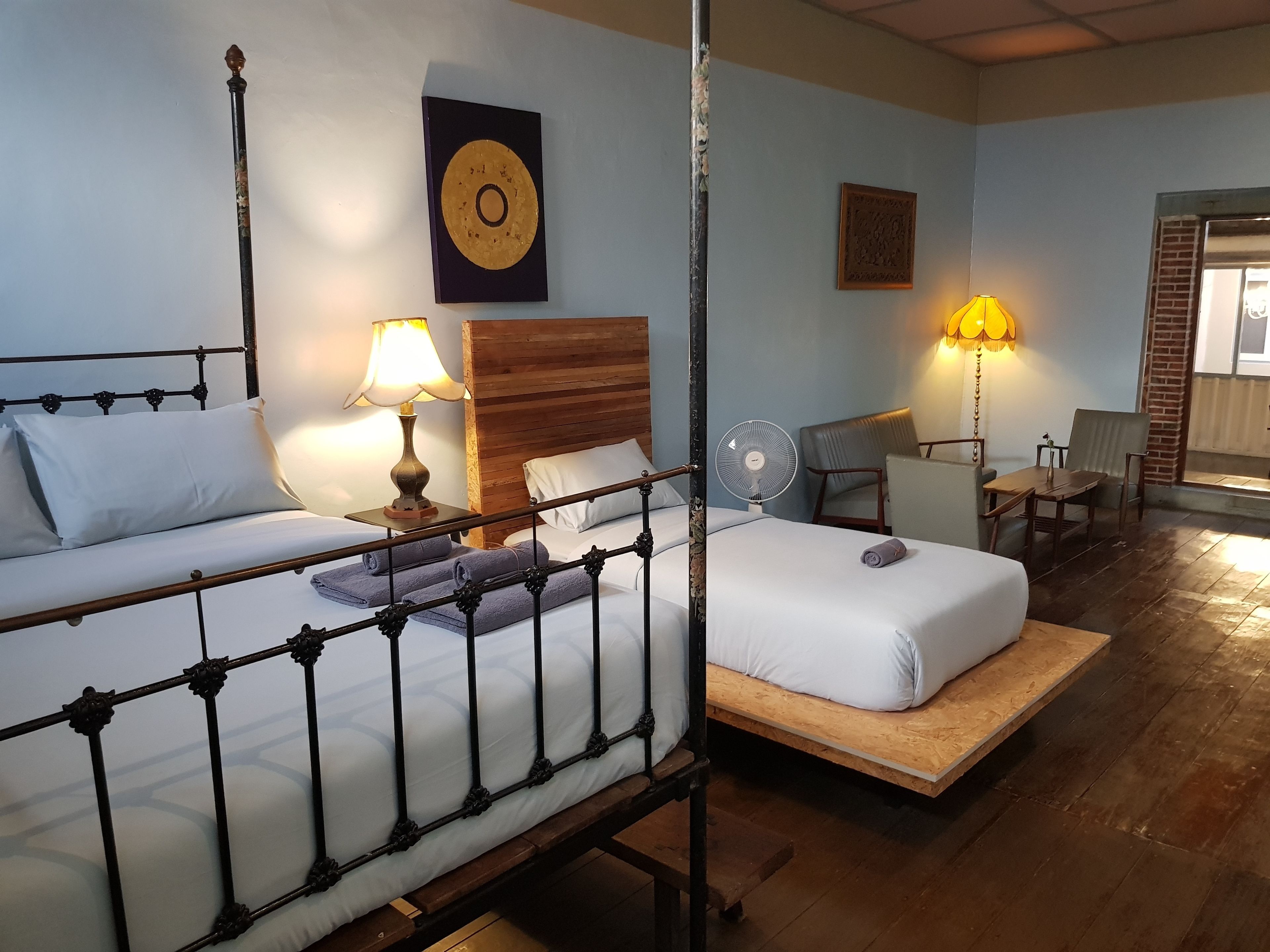 Een hotelkamer met twee bedden, een houten hoofdbord, een wandklok, een ventilator en een zithoek met stoelen en een tafel.