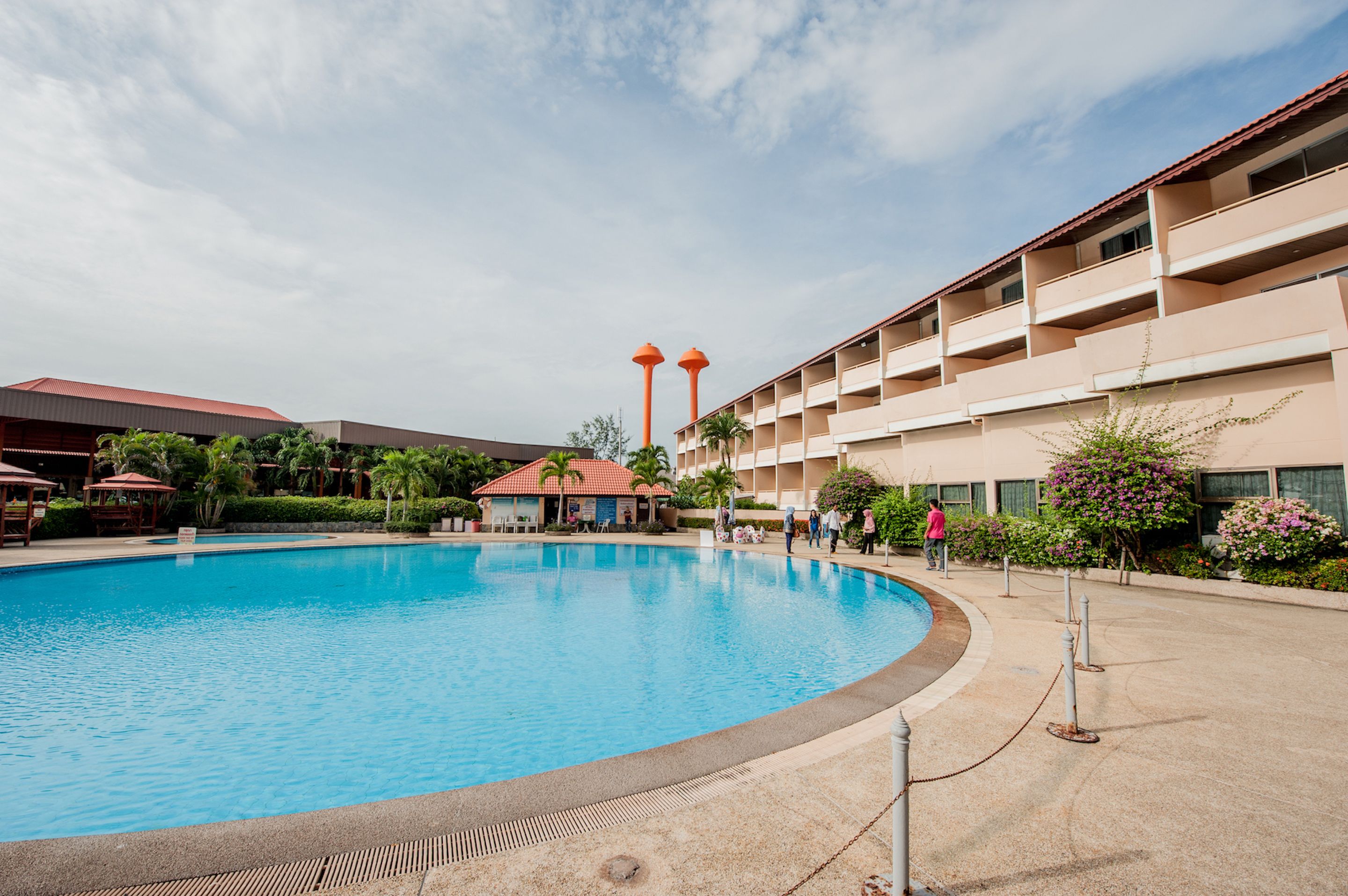A swimming pool surrounded by a hotel building with multiple balconies and a red-roofed structure in the background.