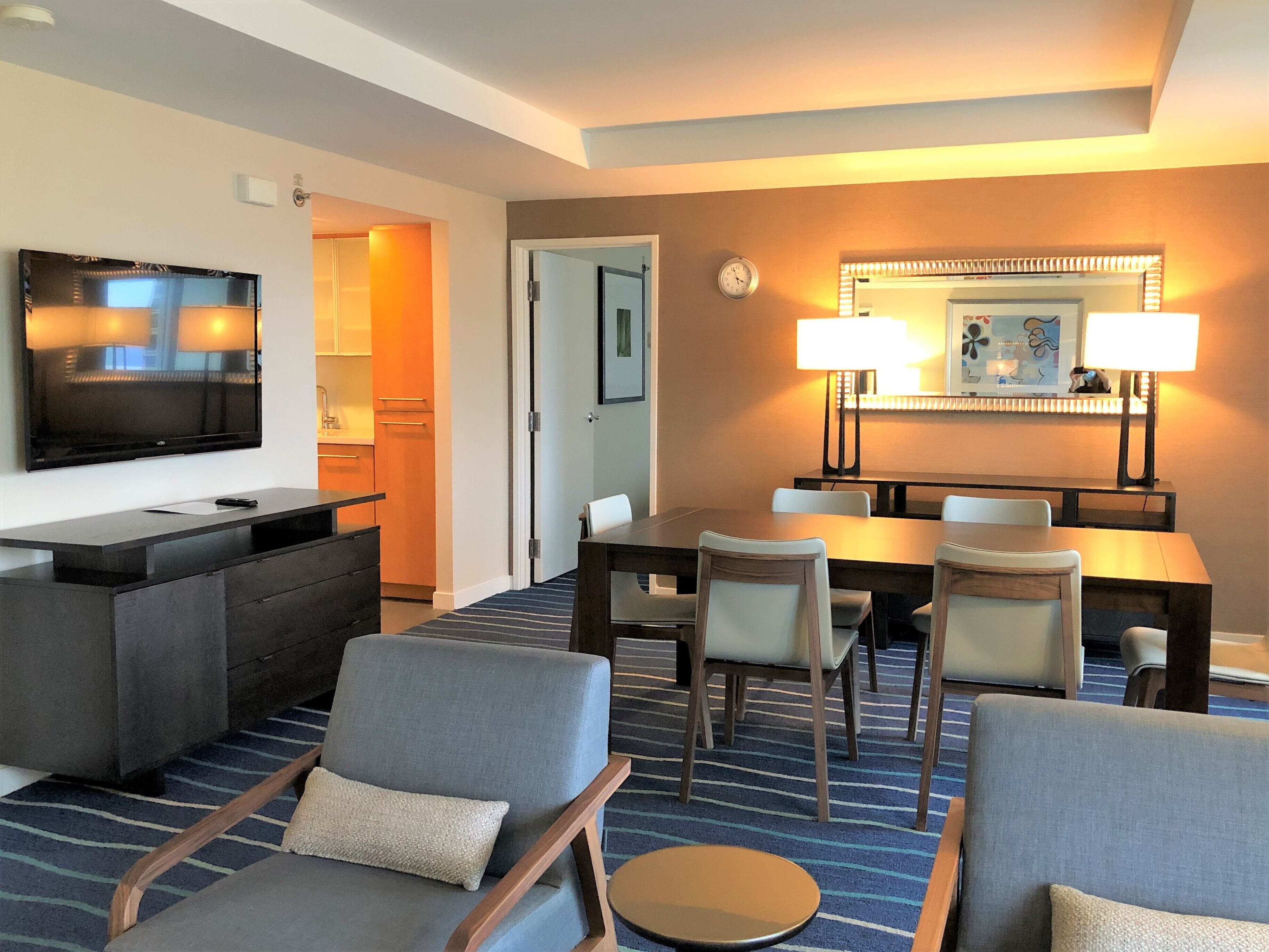 A hotel room with a flat-screen TV, a dining table with chairs, a sofa, and a carpeted floor.