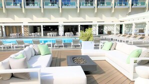 Un espace de détente extérieur, baigné de lumière, avec des canapés blancs et des fauteuils, une piscine, et un bâtiment doté de nombreux balcons en arrière-plan.