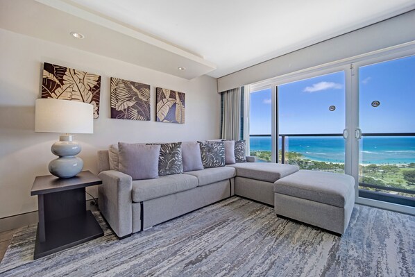 A modern living room with a sectional sofa, a side table with a lamp, and large windows offering a view of the ocean.