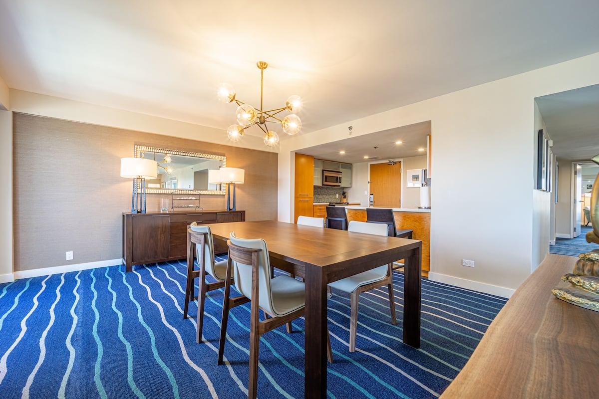 Une chambre d’hôtel dotée d’un coin repas comprenant une table et des chaises en bois, une moquette à motifs et un lustre.